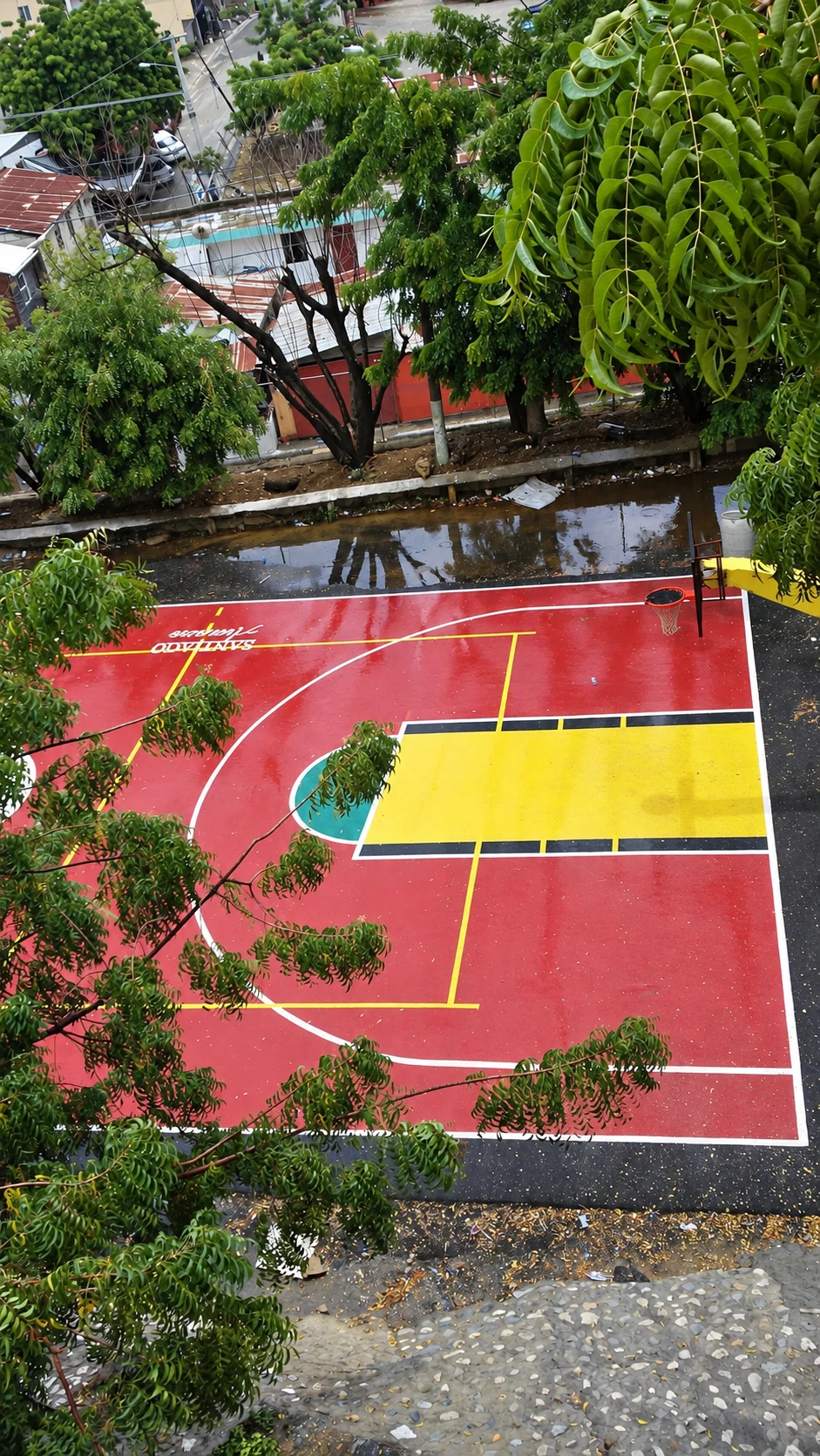 Cancha deportiva vista desde arriba, con pintura roja y amarilla, líneas blancas y árboles alrededor.