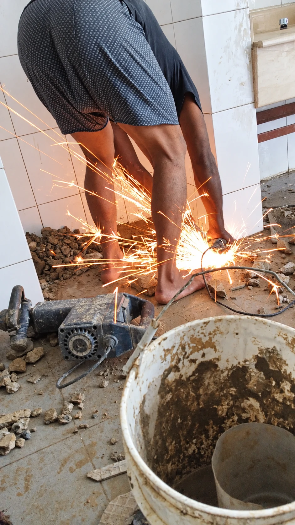 Trabajador inclinado realiza un corte con chispas en un interior con cerámicas, herramientas, escombros y un cubo.
