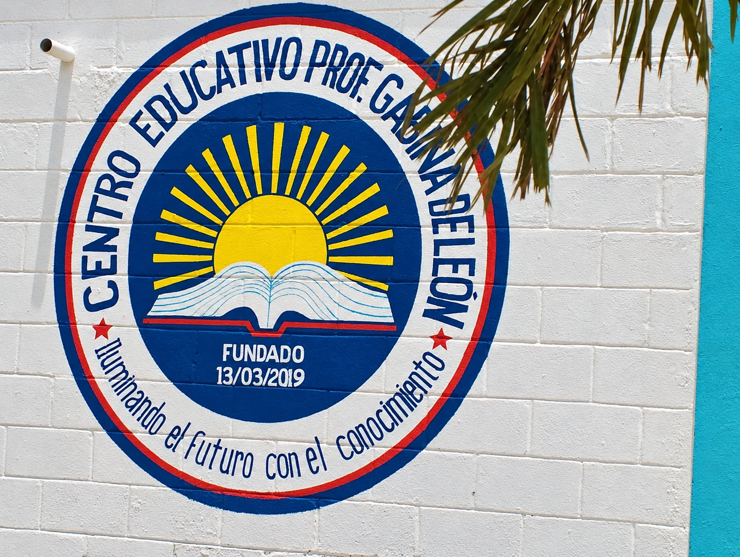 Emblema escolar pintado sobre bloques blancos, con un sol amarillo, un libro y letras azules; unas hojas cubren parte del mural.