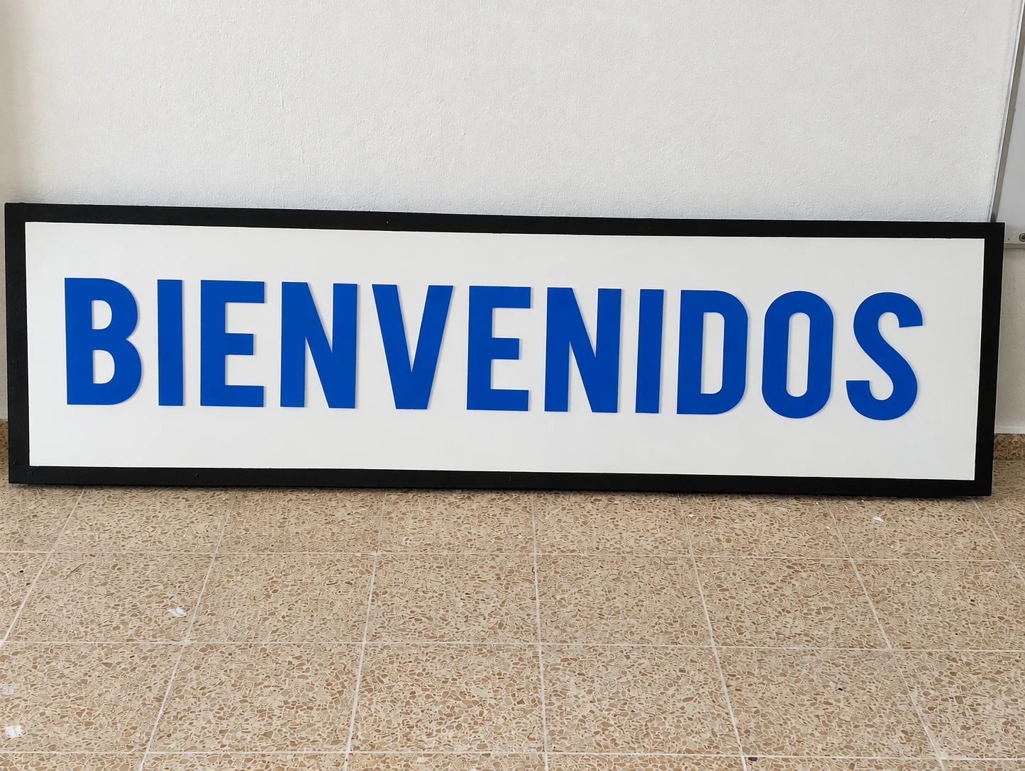 Letrero físico enmarcado en negro con la palabra BIENVENIDOS en letras azules sobre fondo blanco.